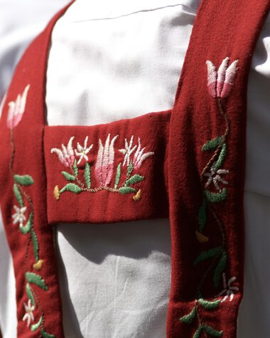 Detailaufnahme einer traditionellen Tracht mit rotem Schulterband, das reichlich bestickt ist, getragen von einer Person in Weiß. | © Kleinwalsertal Tourismus eGen • Fotograf: Hans Wiesenhofer