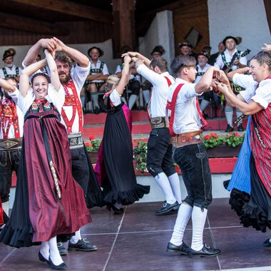 Tanzgruppe in traditionellen Trachten bei einem folkloristischen Fest im Kleinwalsertal, mit Musikern im Hintergrund. | © Kleinwalsertal Tourismus eGen • Fotograf: Frank Drechsel
