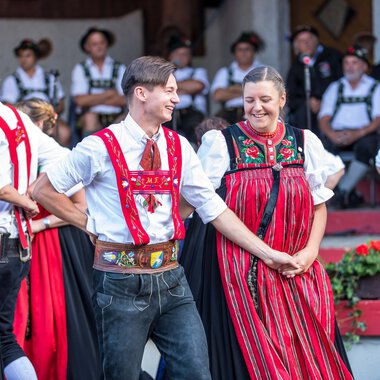 Dancing couple in traditional alpine costumes at an alphorn festival in Kleinwalsertal, surrounded by other dancers in similar attire. | © Kleinwalsertal Tourismus eGen • Photographer: Frank Drechsel