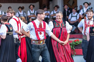 Tanzpaar in traditionellen Alpinskostümen bei einem Alphornfestival in Kleinwalsertal, alle umgeben von weiteren Tanzenden in ähnlicher Tracht. | © Kleinwalsertal Tourismus eGen • Fotograf: Frank Drechsel