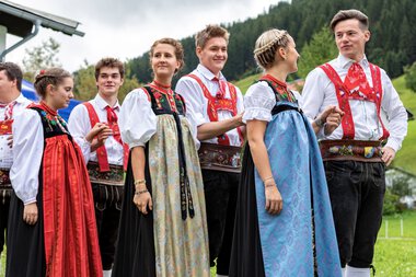 Gruppe von Menschen in traditionellen Trachten beim Alphornfestival im Grünen, mit Bergen im Hintergrund. | © Kleinwalsertal Tourismus eGen • Fotograf: Frank Drechsel