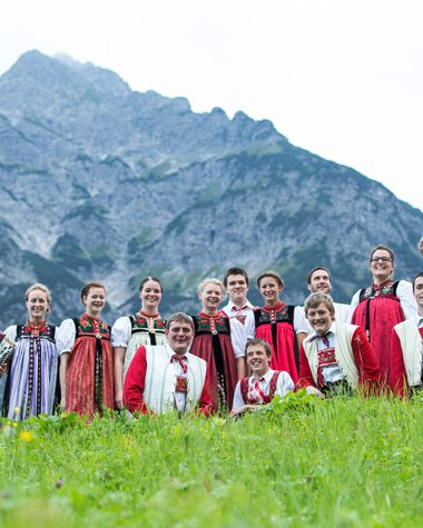 Gruppe von Menschen in traditioneller Kleidung vor einer Bergkulisse, einige mit Musikinstrumenten, im Grünen stehend. | © Kleinwalsertal Tourismus eGen • Fotograf: Frank Drechsel