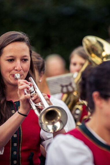 Frau spielt Trompete bei einer Trachtenkapelle im Freien, im Hintergrund andere Musiker und Publikum. | © Kleinwalsertal Tourismus eGen • Fotograf: Oliver Farys