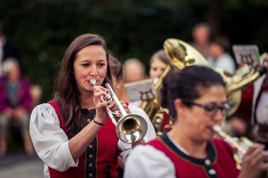 Frau spielt Trompete bei einer Trachtenkapelle im Freien, im Hintergrund andere Musiker und Publikum. | © Kleinwalsertal Tourismus eGen • Fotograf: Oliver Farys
