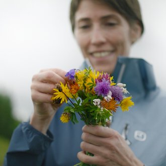 Frau beim Pflücken eines bunten Blumenstraußes im Freien, mit einem Lächeln auf dem Gesicht.