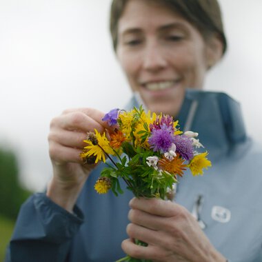 Frau beim Pflücken eines bunten Blumenstraußes im Freien, mit einem Lächeln auf dem Gesicht.