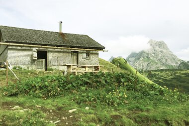 Ein ländliches Haus im Gebirge mit grünen Wiesen, Berge im Hintergrund und bewölktem Himmel.