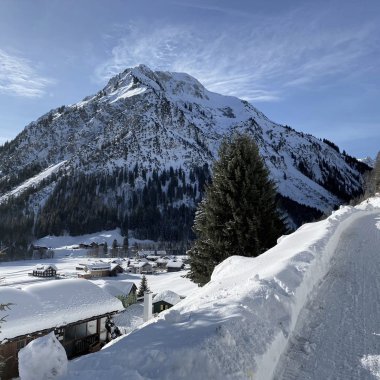 Winterliche Berglandschaft mit schneebedecktem Weg, Häuser und einer großen Bergkulisse im Hintergrund.