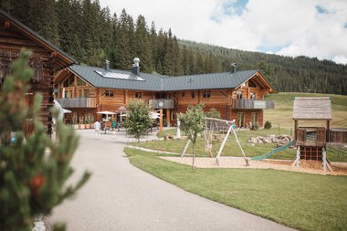 Ein ruhiger Naturzeithof mit Holzgebäuden, Spielplatz und grünen Wiesen inmitten eines Waldes.