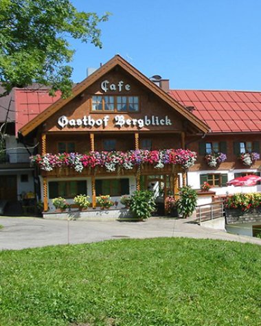 Bergblick Gasthof mit Blumen und Terrasse im Kleinwalsertal, mit einer Ziege im Vordergrund und Hügel im Hintergrund.