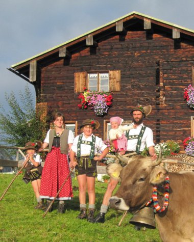Familie in Trachten vor einer Holzalm mit Kühen und Blumen im Kleinwalsertal, Österreich.