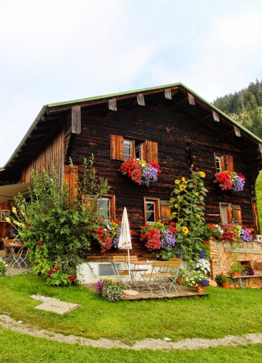 Rustikale, holzgetäfelte Alpenhütte mit bunten Blumen an den Fensterkästen, umgeben von Grünland und mit Bergsicht im Hintergrund.