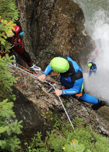 Menschen beim Canyoning in einer engen, steilen Felswand, Wasser stürzt herab, alle tragen Helme und Schutzkleidung.