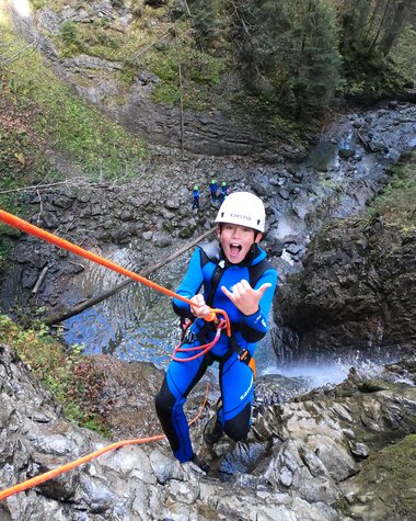 Junge Person beim Canyoning-Abenteuer in einer Naturwasserfalllandschaft, trägt Schutzhelm und Wetsuit.