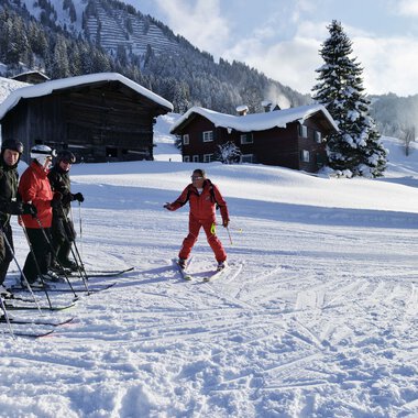 Skilehrer und Teilnehmer beim Skikurs im verschneiten Gebirge, Lehrer zeigt eine Technik, im Hintergrund Hütten und verschneite Bäume.