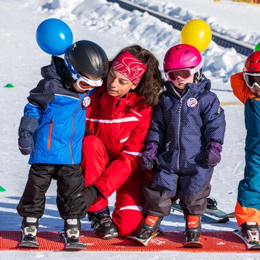 Kinder beim Skischuh-Anziehen im Schnee, mit Skischule-Lehrerin auf einem Wintersportplatz.