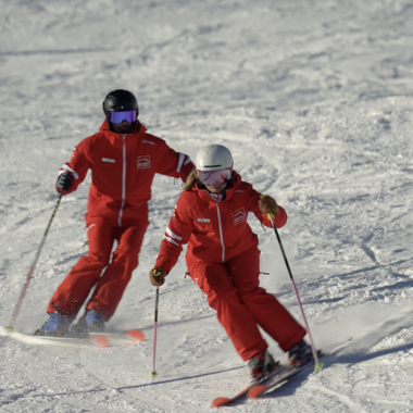 Zwei Skischüler in roten Anzügen beim Skikurs auf Schnee, einer mit Helm und der andere mit Mütze, beim Skifahren an einem verschneiten Hang. | © Kleinwalsertal Tourismus | Skischule Hirschegg