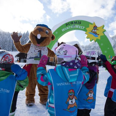 Kinder beim Skikurs in Riezlern begrüßen den Burmi-Winterwelten-Maskottchen vor einem Winterhintergrund mit Bergen.