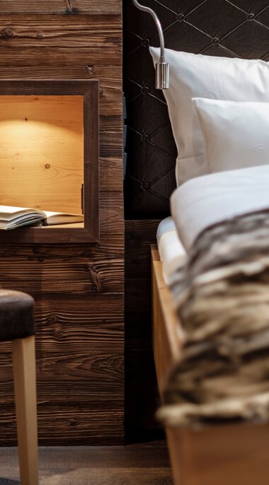 Innenansicht eines gemütlichen Zimmers mit einem Bett, Kopfteil in Schwarz, und einer Holzbox an der Wand, die ein Buch enthält.