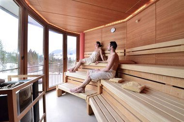 Zwei Personen sitzen in einem Saunabereich mit Holzregalen und großen Fenstern, die eine verschneite Landschaft zeigen.