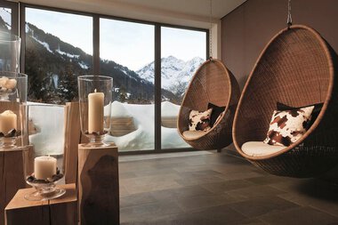 Gemütlicher Raum im Ifen Travelcharme Hotel mit Blick auf verschneite Berge durch große Fenster, zwei Hängesessel und Kerzen.