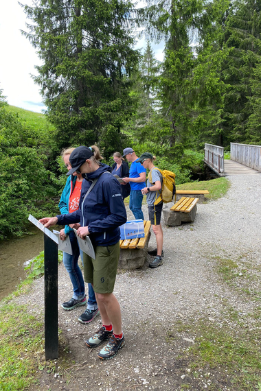 Gruppe von Menschen liest Informationen an einer Infotafel in einer grünen Waldlandschaft beim Wandern.