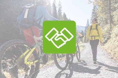 Zwei Fahrradfahrer und ein Wanderer auf einem Waldweg, bei Sonnenschein, umgeben von Bäumen.