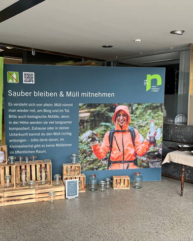 Informationsstand zur Müllkampagne, mit einem großen blau-grünen Poster, Holzboxen, und zwei Tischen mit Tablets, im Innenraum.
