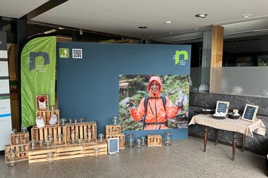 Ausstellung mit Informationsständen und einem Poster einer Person in orangefarbener Jacke, im Hintergrund grüne Dekoration. | © Kleinwalsertal Tourismus eGen