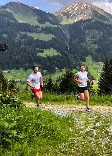 Zwei Menschen laufen auf einem Waldweg in den Bergen mit grünen Wiesen und hohen Gipfeln im Hintergrund. | © Kleinwalsertal Tourismus eGen 