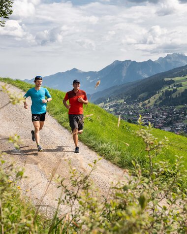 Zwei Läufer beim Trailrunning in den Bergen, unterwegs auf einer schmalen Wanderweg im Grünen mit горigem Panorama im Hintergrund. | © Kleinwalsertal Tourismus eGen • Fotograf: Rupert Mühlbacher