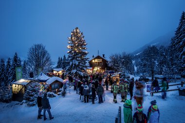 Festliche Winterveranstaltung am Adventszauber mit Besuchern, Weihnachtsbäumen und festlichem Lichterglanz in einer verschneiten Landschaft.