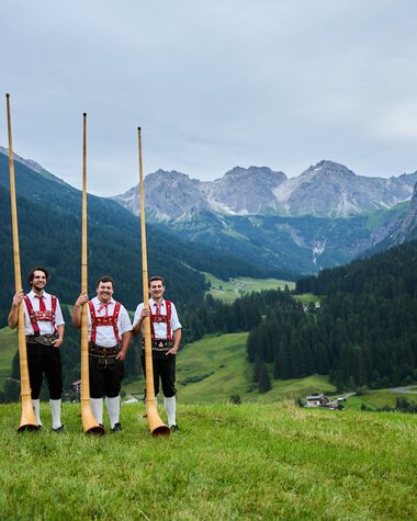 Drei Männer in Trachten stehen in einer grünen Wiese mit Alphörnern, umgeben von Bergen und einer bewaldeten Talregion.