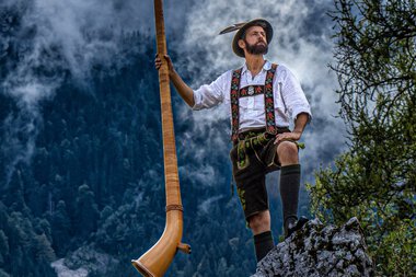 Man in traditional attire with an Alphorn on a rock in the mountains, looking upward, with clouds and trees in the background. | © Loisach Marci • Anton Ostler
