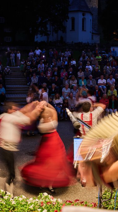Musiker und Tänzer beim Alphornfestival im Kleinwalsertal, im Hintergrund ein großer Publikum im Freien bei Abenddämmerung.