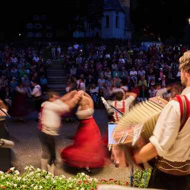 Musiker und Tänzer beim Alphornfestival im Kleinwalsertal, im Hintergrund ein großer Publikum im Freien bei Abenddämmerung.
