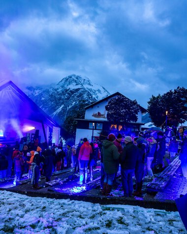 People gather at an Alpine horn festival in a snowy mountain setting during nighttime with blue lighting.