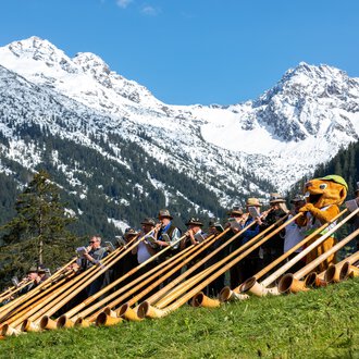 Gruppenfoto beim Alphornfestival 2024 mit Musikern in traditioneller Kleidung und Alphörnern, vor atemberaubender Bergkulisse mit schneebedeckten Gipfeln.