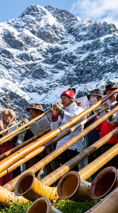 People at the alphorn festival in the mountains, with a mountain peak in the background, playing traditional alphorn instruments. | © Kleinwalsertal Tourismus eGen • Photographer: Frank Drechsel