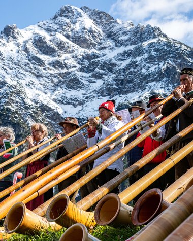 Menschen beim Alphornfestival in den Bergen, mit Berggipfel im Hintergrund, spielen auf traditionellen Alphorninstrumenten.