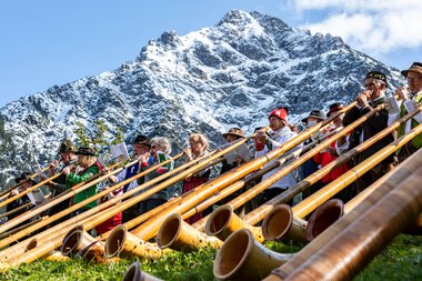 Musiker beim Alphornfestival vor beeindruckender Bergkulisse, mit vielen Alphörnern auf dem Gras liegend.