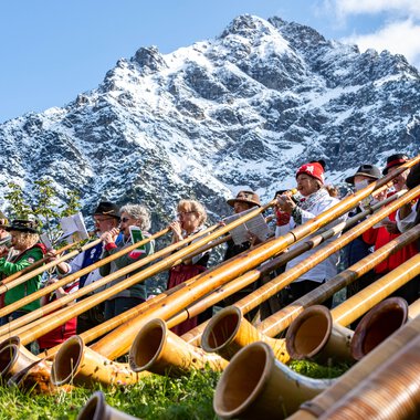 Musiker beim Alphornfestival vor beeindruckender Bergkulisse, mit vielen Alphörnern auf dem Gras liegend.