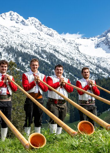 Fünf Männer spielen Alphorn vor einer Bergkulisse mit Schnee bedeckten Gipfeln und grünen Wäldern. | © Kleinwalsertal Tourismus eGen • Fotograf: Frank Drechsel