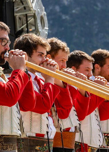 Fünf Männer in traditioneller Tracht spielen auf Alphörnern bei einem Fest im Freien.