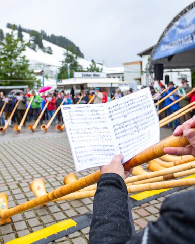 Orchester bei einem Alphornfestival, Musiker spielen das Alphorn, während das Publikum im Freien bei Regen zuschaut. | © Kleinwalsertal Tourismus eGen • Fotograf: Frank Drechsel