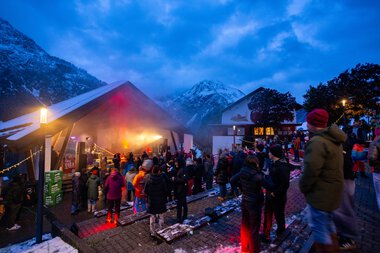 Menschen versammeln sich beim Alphornfestival 2024, das in einer Bergregion bei Dämmerung stattfindet, mit einer Bühne im Freien und schneebedeckten Bergen im Hintergrund.
