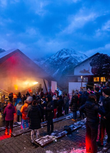 People gather at the Alphorn Festival 2024 in a mountain area at dusk, with an outdoor stage and snow-capped peaks in the background.