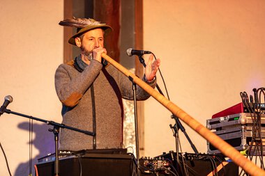 Ein Musiker spielt das Alphorn bei einer Veranstaltung, ausgestattet mit traditionellem Hut und moderner Ausrüstung im Hintergrund.