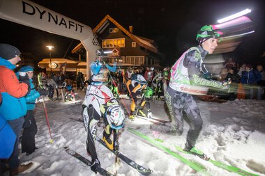 Ski- und Skitourenrennen bei Nacht vor einem gemütlichen Haus, mit mehreren Teilnehmern und Zuschauern im Schnee.