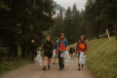 Menschen nehmen an einer baumpflege- oder sampelaktion in einem Wald teil, tragen Müllsäcke und gehen den Weg entlang. | © Patron e.V. • Fotograf: Michelle Meitzner